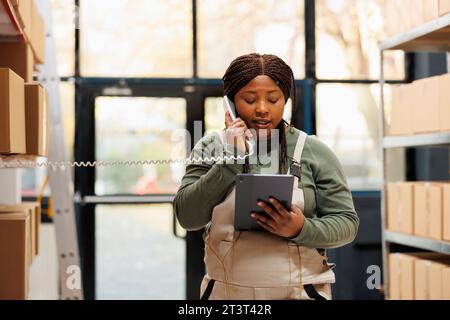 Responsable répondant à un appel téléphonique fixe dans l'entrepôt, discutant avec le superviseur à distance du contrôle qualité des produits. Travailleur afro-américain portant l'ensemble tout en vérifiant la logistique sur tablette Banque D'Images
