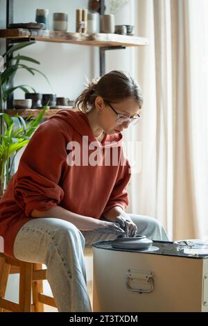 Femme détendue faisant des ustensiles en argile naturelle s'asseoir dans l'atelier à la table avec support de filature Banque D'Images