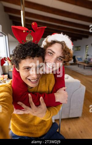 Portrait d'heureux couple masculin gay diversifié portant des chapeaux de noël et ayant un appel vidéo à la maison Banque D'Images