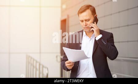 Homme d'affaires concentré en costume parle sur le téléphone tenant des papiers Banque D'Images