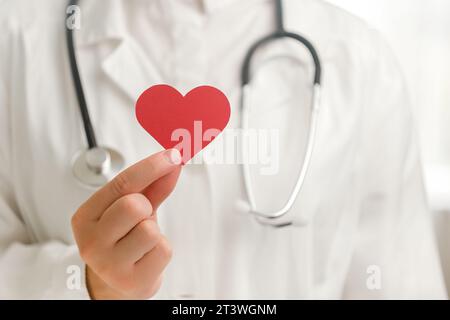 Gros plan d'une femme inconnue médecin en uniforme blanc avec stéthoscope tenant une petite forme de coeur rouge à l'hôpital. Amour, donneur, journée mondiale du cœur, monde guérit Banque D'Images