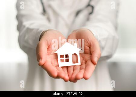 Foyer sélectif de jeune médecin inconnu portant l'uniforme blanc médical professionnel et stéthoscope tenant dans les mains petite maison de papier, debout Banque D'Images