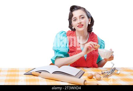 Belle femme au foyer de style vintage préparant un dessert à la maison, elle vérifie la recette dans un livre de cuisine et mélange les ingrédients dans un bol Banque D'Images