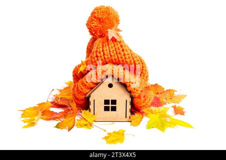 Charmante maison miniature en bois portant un chapeau en tricot orange chaud orné de feuilles d'automne isolé sur blanc. Confort et chaleur en saison froide c Banque D'Images