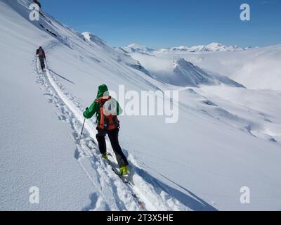 Deux personnes skiant dans l'arrière-pays sur une piste à travers la neige par une journée ensoleillée. Banque D'Images