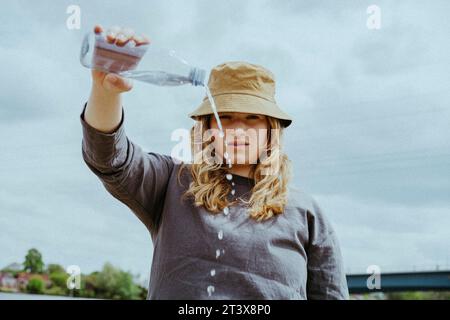 Portrait d'angle bas de jeune femme versant de l'eau de bouteille contre le ciel Banque D'Images