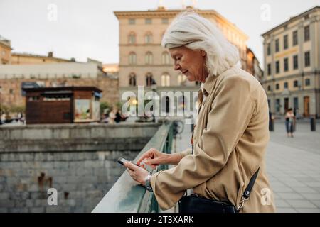 Vue latérale de la femme âgée appuyée sur la balustrade tout en utilisant un téléphone intelligent en ville Banque D'Images