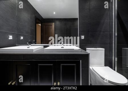 Image frontale de salle de bains carrelée avec des carreaux d'ardoise noire, toilettes blanches, lavabos individuels en porcelaine blanche sur armoire noire avec miroir intégré dans la wa Banque D'Images