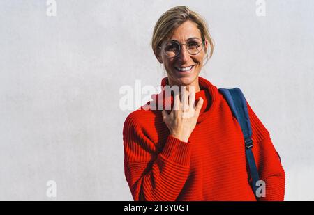 Portrait d'une jeune femme au sourire agréable et aux lunettes qui saisit son col roulé rouge isolé sur un mur de béton avec espace copie. Belle étudiante Banque D'Images