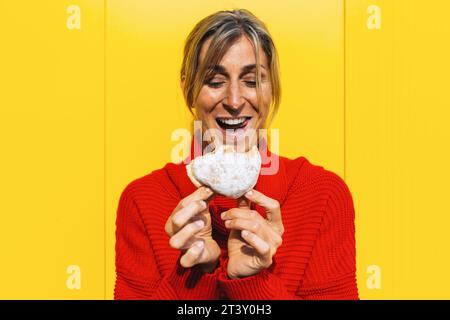 Femme enthousiaste mordre dans un beignet berliner rempli de confiture de fraises contre le mur jaune Banque D'Images