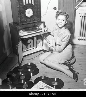 Femme au gramophone dans les années 1940 Elle a beaucoup de disques différents à choisir pour écouter. Une radio est sur le dessus du gramophone. Les disques étaient faits d'un matériau fragile et la vitesse du disque à sonner comme il se doit était de 78 tours par minute. Notez les petites boîtes en fer blanc à côté du gramophone. Ils contiennent les aiguilles qui sont le support pour lire les enregistrements avec, l'aiguille courant dans les lignes fines de l'enregistrement. Les aiguilles ont dû être remplacées car elles étaient usées, et vous avez fait cela vous-même. Suède 1945. Kristoffersson réf. O124-5 Banque D'Images