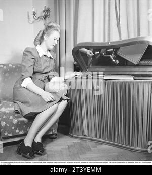 Femme au gramophone dans les années 1940 Elle a mis un disque et écoute la musique. Les disques étaient faits d'un matériau fragile et la vitesse du disque à sonner comme il se doit était de 78 tours par minute. Le gramophone est un modèle de luxe avec un tissu satiné couvrant les côtés. Suède 1947. Kristoffersson réf. AB22-2 Banque D'Images