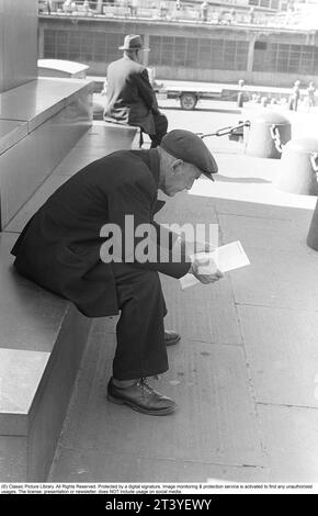 Dans les années 1950 Un homme âgé assis sur les marches lisant la liste des chevaux de départ aux prochains hippodromes sur la piste de course Solvalla. Il est vêtu d'une veste et d'une casquette et a l'air réfléchi lorsqu'il parcourt la liste des chevaux de départ pour choisir le gagnant. Stockholm Suède 1953 ref DV1 Banque D'Images