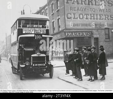 Jalon dans l'histoire des bus britanniques. Le 2 octobre 1925, le premier voyage en bus se déroule avec un bus à impériale dont l'étage supérieur est recouvert d'un toit. Auparavant, les bus n'avaient pas de toit. C'est le bus numéro 100 qui relie Epping Town à Elephant and Castle, et les gens font la queue pour embarquer à Leytonstone dans l'est de Londres où le bus s'arrête lors de sa première visite. Le bus a 10 fenêtres sur le nouvel étage supérieur couvert, au total il y avait de la place pour 52 passagers assis. Le bus est du modèle LGOC qui a été introduit à l'origine à Londres en 1910 et a été construit et exploité par le London Gen Banque D'Images