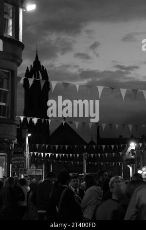 Photo de portrait en échelle de gris de la tour de l'église et des bannières pendant le festival Celtic Beltane la nuit avec des gens vus en train de moudre ci-dessous (Peebles, Écosse) Banque D'Images