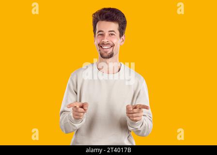 Beau jeune homme aux cheveux épais et à la barbe sourit, pointant ses index vers vous, devant lui vers la caméra. Banque D'Images