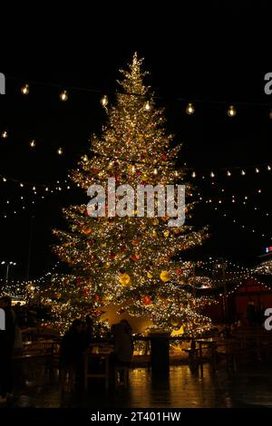 Vrai, entièrement décoré avec des ornements ou des boules d'or et rouge, arbre de vacances d'hiver de Noël enfilé avec des lumières de fée d'or jaune la nuit dans un marché Banque D'Images