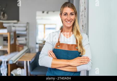 femme blonde joyeuse avec les bras croisés en tablier se tient dans un espace de travail de tailleur. Elle a un ruban à mesurer et des ciseaux dans sa poche de tablier et est sur Banque D'Images