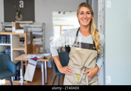 femme blonde joyeuse dans un haut gris et tablier beige se tient dans un espace de travail de tailleur. Elle a un ruban à mesurer et des ciseaux dans sa poche de tablier et est s. Banque D'Images