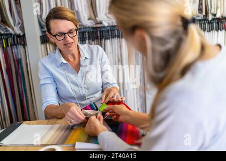 Couturière est assise avec son client dans la boutique et la conseille sur le choix de son tissu de rideau. Couturière souriant et touchant un échantillon de couleur tandis que th Banque D'Images