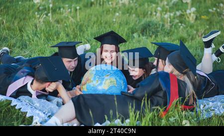 Diplômés en robes noires regardant un globe géorgraphique allongé sur l'herbe. Banque D'Images