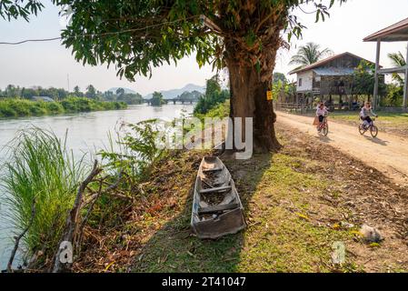 Février 22 2023 : une scène rurale paisible, comme les villageois pédalent le long du chemin de terre, courant à côté de la rivière, devant un arbre feuillu et vieux bateau de rivière, sur un ho Banque D'Images