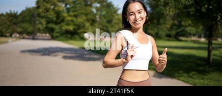 Une fille de forme physique souriante montre le pouce vers le haut, se tient avec une serviette après une bonne séance d'entraînement dans le parc, le jogging et l'air heureux Banque D'Images