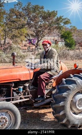 vieil agriculteur africain conduisant un tracteur sur le champ devant le village Banque D'Images