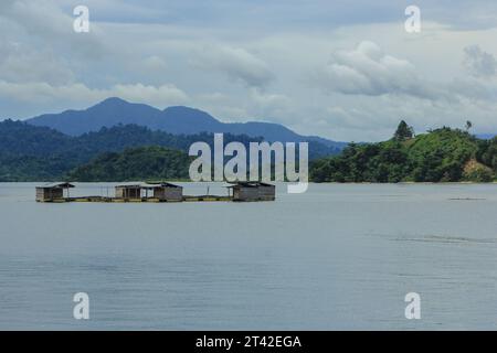 Village flottant isolé avec des maisons en bois sur pilotis sur un lac serein, entouré de montagnes verdoyantes sous un ciel nuageux spectaculaire. Banque D'Images