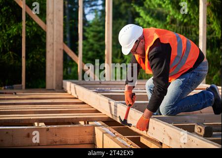 Carpenter fabrique une maison à ossature en bois. Un homme barbu portant des lunettes cloue avec un marteau, vêtu d'un casque de protection et d'un gilet de construction. Banque D'Images