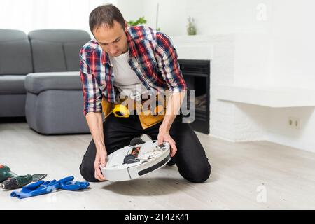 Un jeune homme de race blanche effectue la maintenance d'un robot-aspirateur blanc. Aspirateur moderne sans fil autonome. Nettoyant robot de réparation pour homme à la maison Banque D'Images