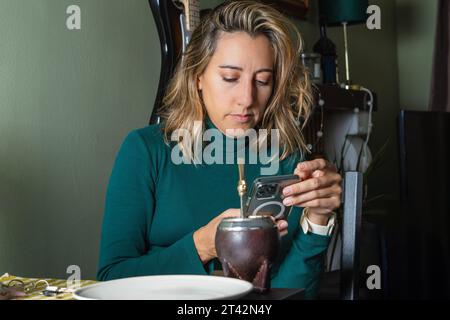 Une jeune femme portant une robe blanche tient un verre de vin dans sa main droite tout en regardant son téléphone portable avec sa main gauche sur une table Banque D'Images