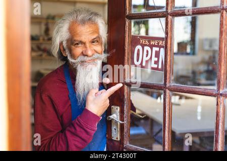 Heureux propriétaire d'entreprise biracial senior avec une longue barbe montrant le signe de magasin à ouvrir Banque D'Images