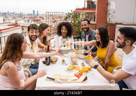 Groupe joyeux d'amis divers mangeant et buvant à une table de terrasse. Des gens jeunes et détendus s'amusant dans un dîner sur le toit. Banque D'Images