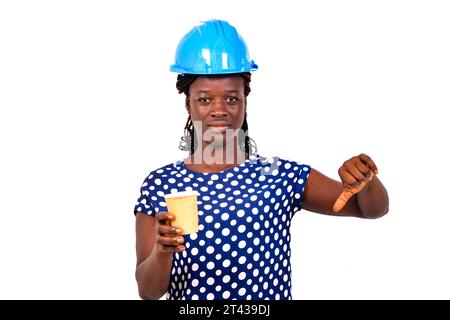 belle jeune femme ingénieur en construction portant un casque de sécurité bleu tenant une tasse à café en papier et pointant son pouce vers le bas. Banque D'Images