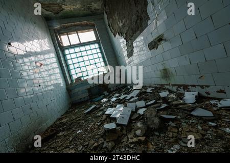 D'en haut de la salle de mur de carreaux de céramique réfléchissant dans la lumière du jour tandis que les gravats éparpillés brisés en tas sur le sol près de la fenêtre avec la clôture et le plafond de ciment s'effritant, de place abandonnée Banque D'Images