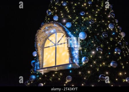 Une nuit d'hiver enchanteresse : un grand sapin de Noël se dresse haut, projetant une lueur chaude à travers une fenêtre illuminée, évoquant la magie des fêtes Banque D'Images