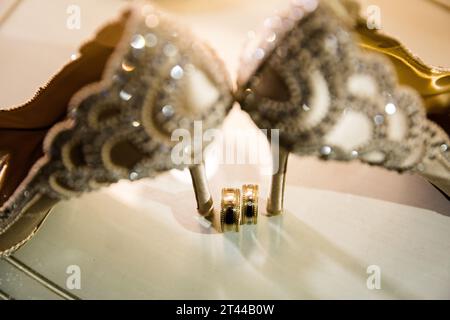 Élégants talons de mariée blancs et bague de mariage sur une surface réfléchissante, avec un décor de pièce luxueusement flouté. Banque D'Images