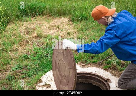 Un plombier ouvre un couvercle de trou d'homme sur un puits de béton. Inspection et entretien des puits d'eau et d'égout. Banque D'Images