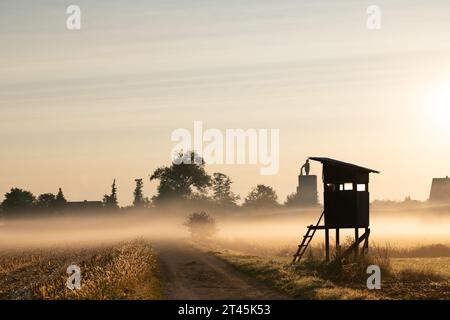 Un stand de chasse en bois se dresse dans un champ ouvert. Le soleil se lève. Il y a du brouillard et de la brume au-dessus du champ. Le paysage est baigné de lumière rouge. Arbres et h Banque D'Images