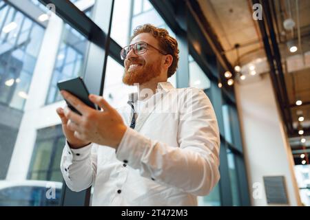 Freelance homme barbu tenant le téléphone tout en se tenant au bureau pendant le temps de pause et regarde loin Banque D'Images