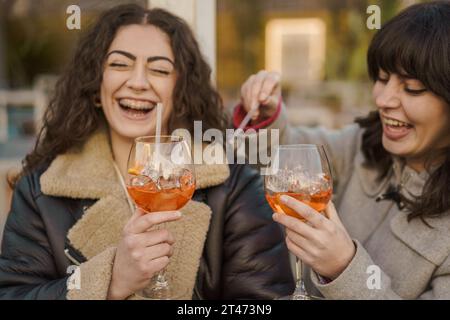 Deux femmes rayonnent de bonheur en dégustant leurs cocktails spritz en plein air, capturant un moment spontané de joie et d’amitié. Banque D'Images