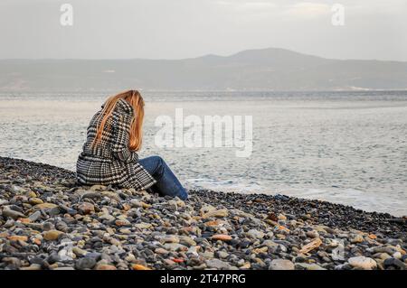 Une femme blonde assise au bord de la mer sur une plage de galets en automne Banque D'Images
