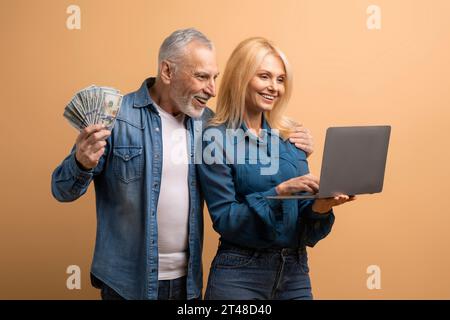 Couple senior émotionnel utilisant un ordinateur portable sur fond beige, montrant de l'argent Banque D'Images
