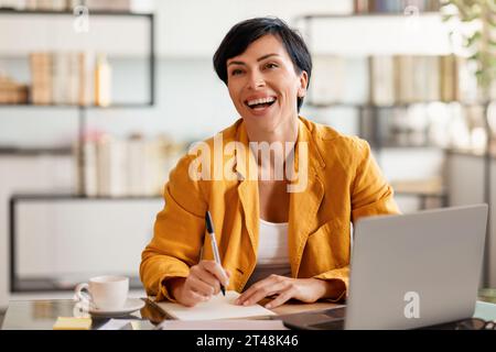 Heureuse femme d'affaires brunette d'âge moyen écrivant des papiers assis au bureau Banque D'Images