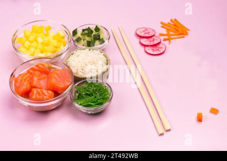 Ensemble de légumes crus, poisson rouge, riz, sauce soja dans des bols en verre. Plat hawaïen. Baguettes de bambou sur fond rose. Vue de dessus Banque D'Images