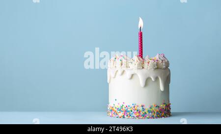 Gâteau d'anniversaire de célébration décoré avec glaçage goutte à goutte blanc, tourbillons de glaçage à la crème au beurre, saupoudrages de sucre colorés et une bougie d'anniversaire contre un pla Banque D'Images