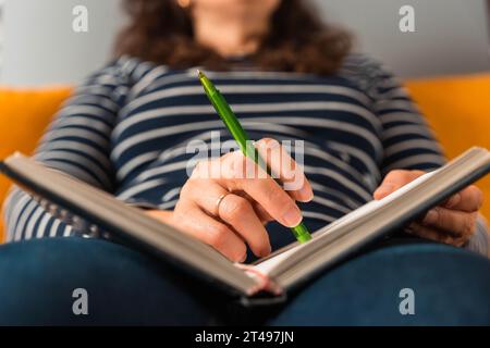 femme méconnaissable écrit des notes avec un stylo dans un bloc-notes. Elle pensa en étant assise sur le canapé. Son objectif est d'attraper chaque pensée, inspi Banque D'Images