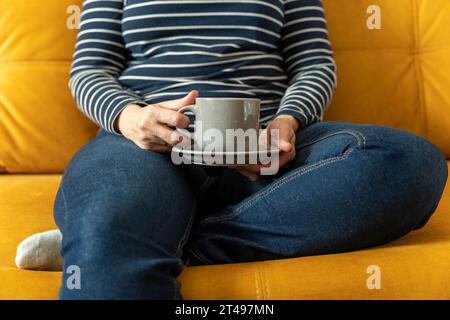 Une femme méconnaissable dans un pull rayé et un Jean bleu s'assoit sur le canapé avec sa jambe cachée sous elle et tient une tasse de café dans ses mains. gir Banque D'Images