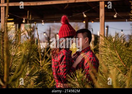 Tendres câlins de jeune couple amoureux parmi le marché vert des arbres de Noël en ville. Belle femme embrasse passionnément, regarde l'homme. Les jeunes mariés regardent Banque D'Images